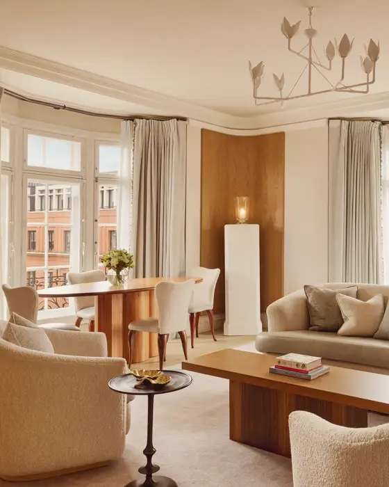 Bright and elegant living room with curved cream sofas, wooden coffee table, and dining area by tall bay windows framed with soft grey curtains overlooking red-brick buildings.