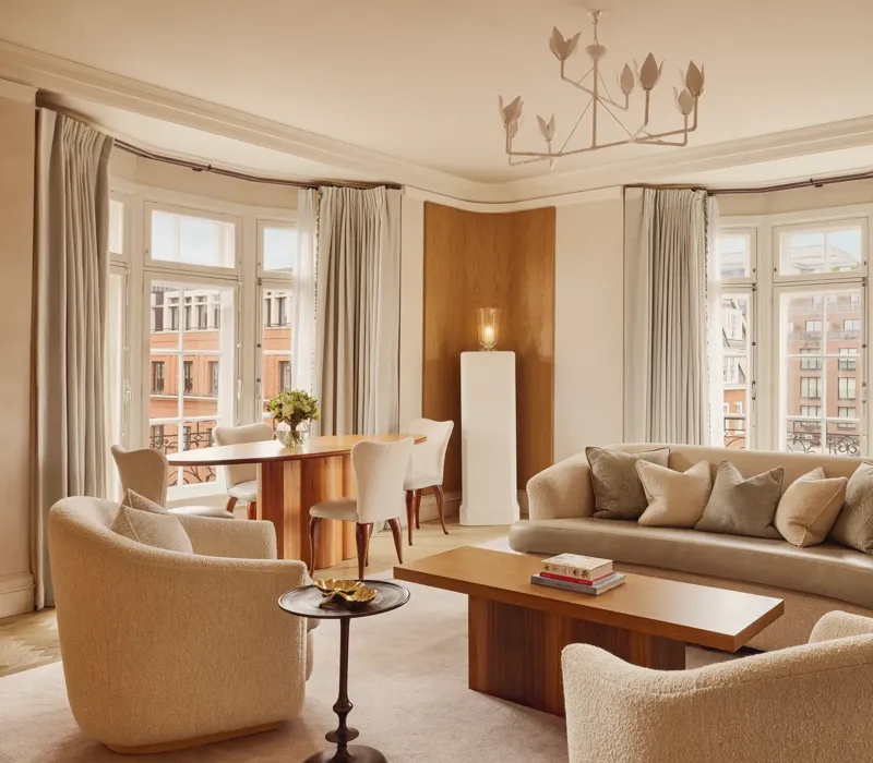Bright and elegant living room with curved cream sofas, wooden coffee table, and dining area by tall bay windows framed with soft grey curtains overlooking red-brick buildings.