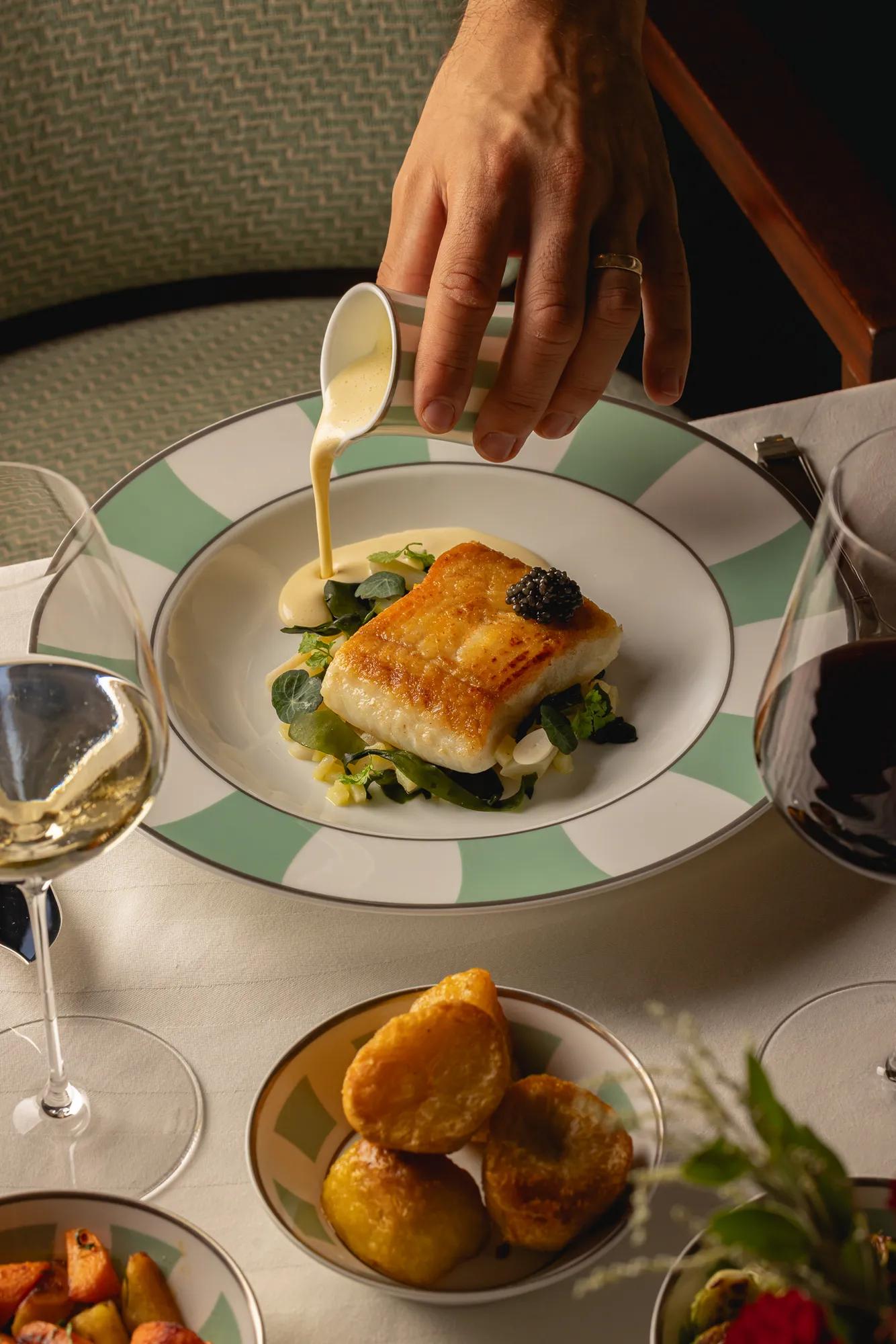 Seared white fish fillet topped with caviar, served on greens in creamy sauce on striped rim plate.