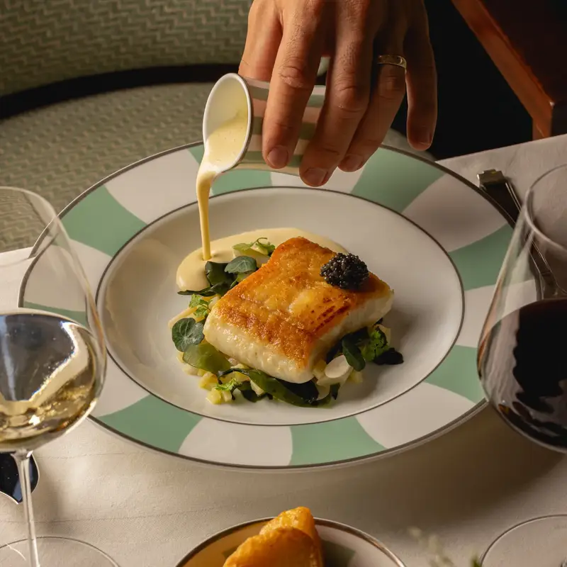 Seared white fish fillet topped with caviar, served on greens in creamy sauce on striped rim plate.