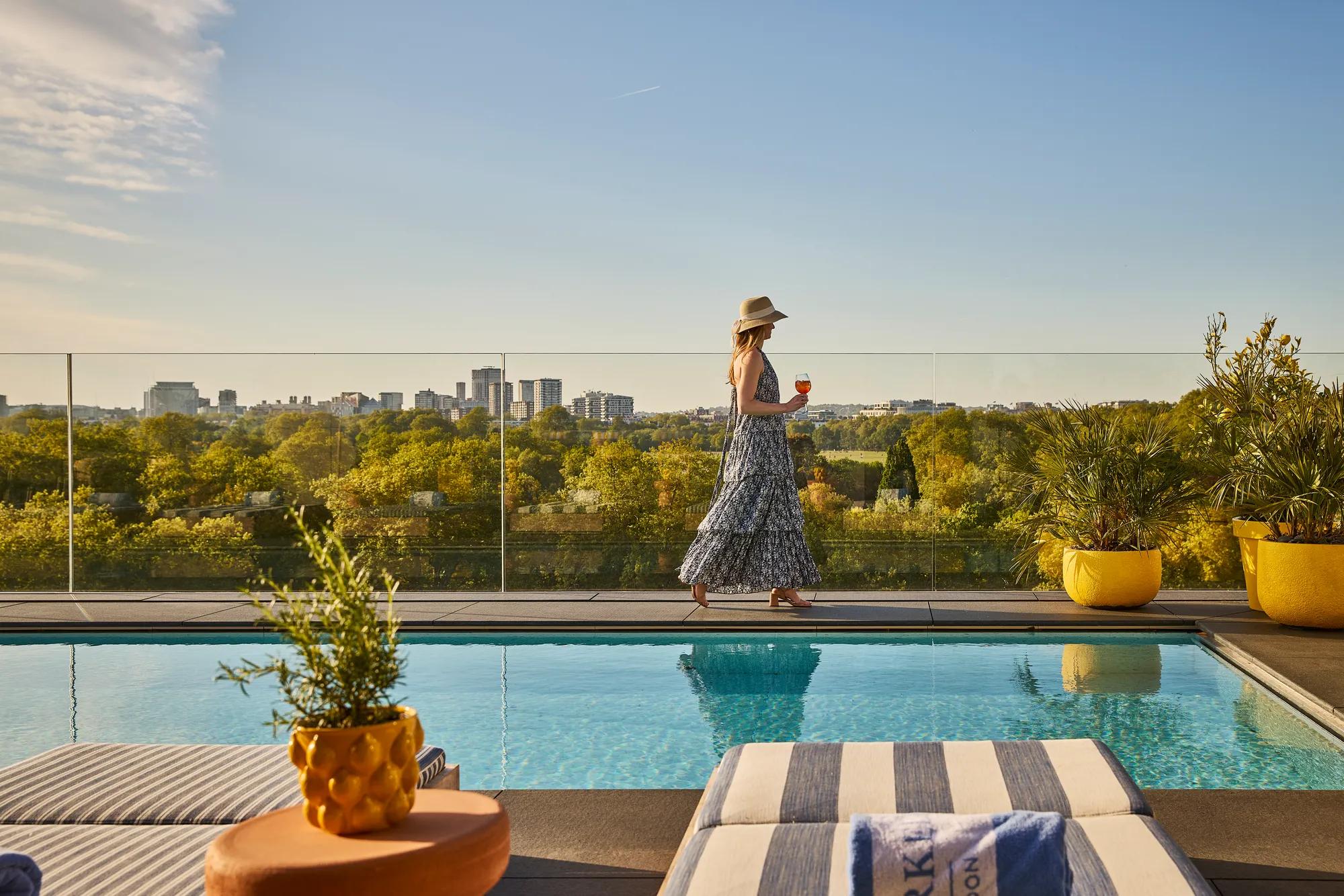Guest walking beside a rooftop pool with striped loungers, glass railings and sweeping views across the park and city skyline.