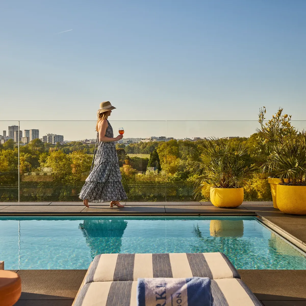Guest walking beside a rooftop pool with striped loungers, glass railings and sweeping views across the park and city skyline.