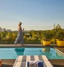 Guest walking beside a rooftop pool with striped loungers, glass railings and sweeping views across the park and city skyline.