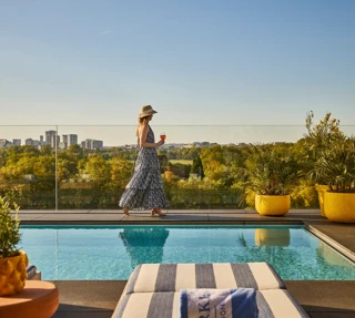 Guest walking beside a rooftop pool with striped loungers, glass railings and sweeping views across the park and city skyline.
