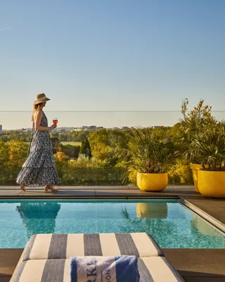 Guest walking beside a rooftop pool with striped loungers, glass railings and sweeping views across the park and city skyline.