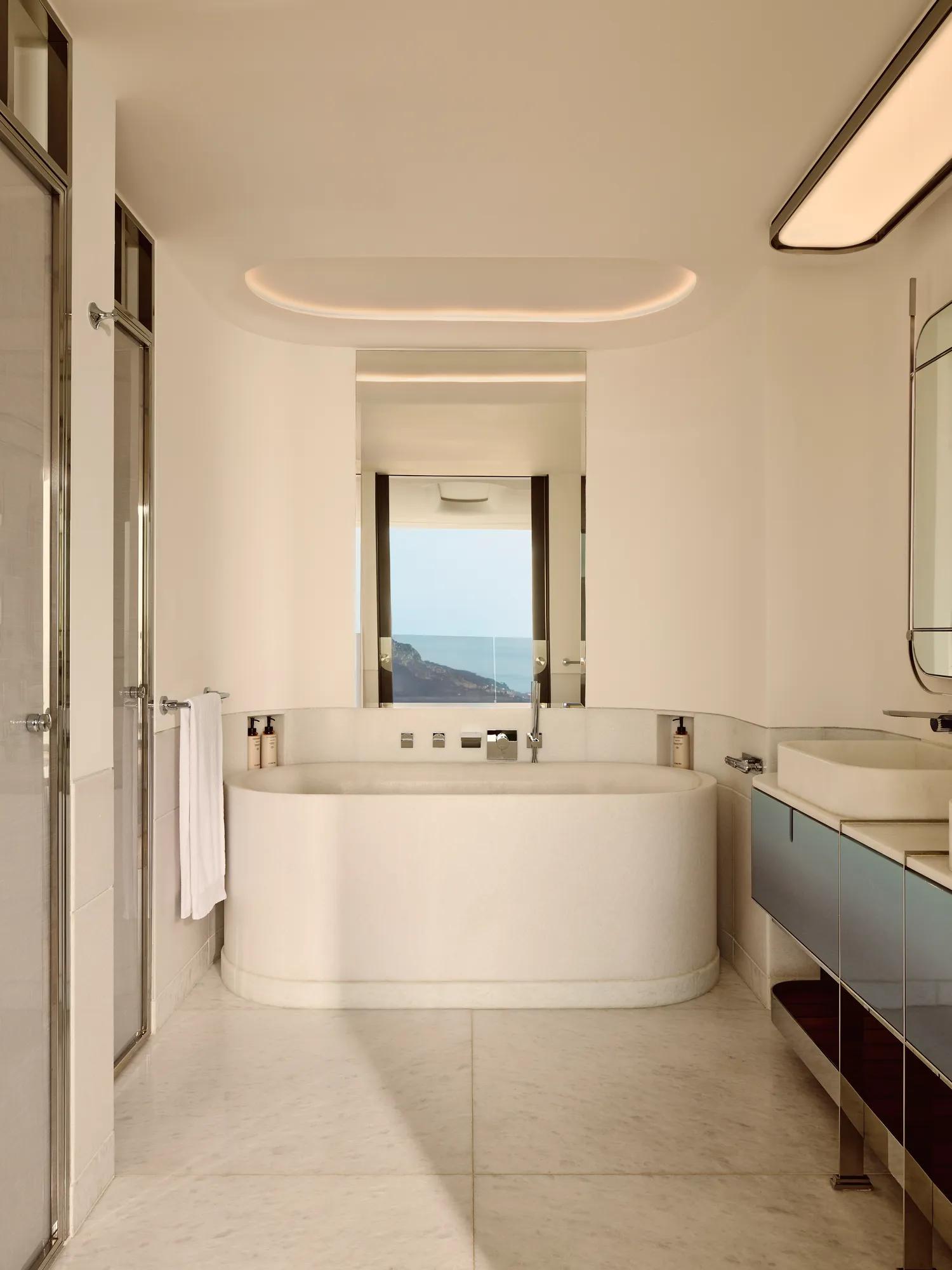Modern bathroom with a freestanding oval tub, marble surfaces, and a window offering a sea view..