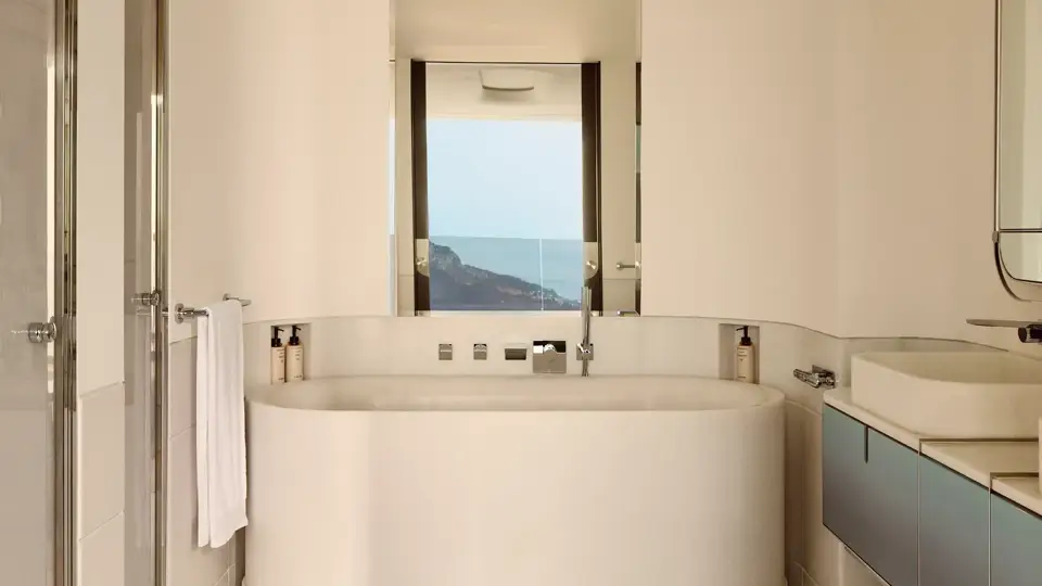 Salle de bain moderne avec baignoire ovale, surfaces en marbre et vue sur la mer par la fenêtre.