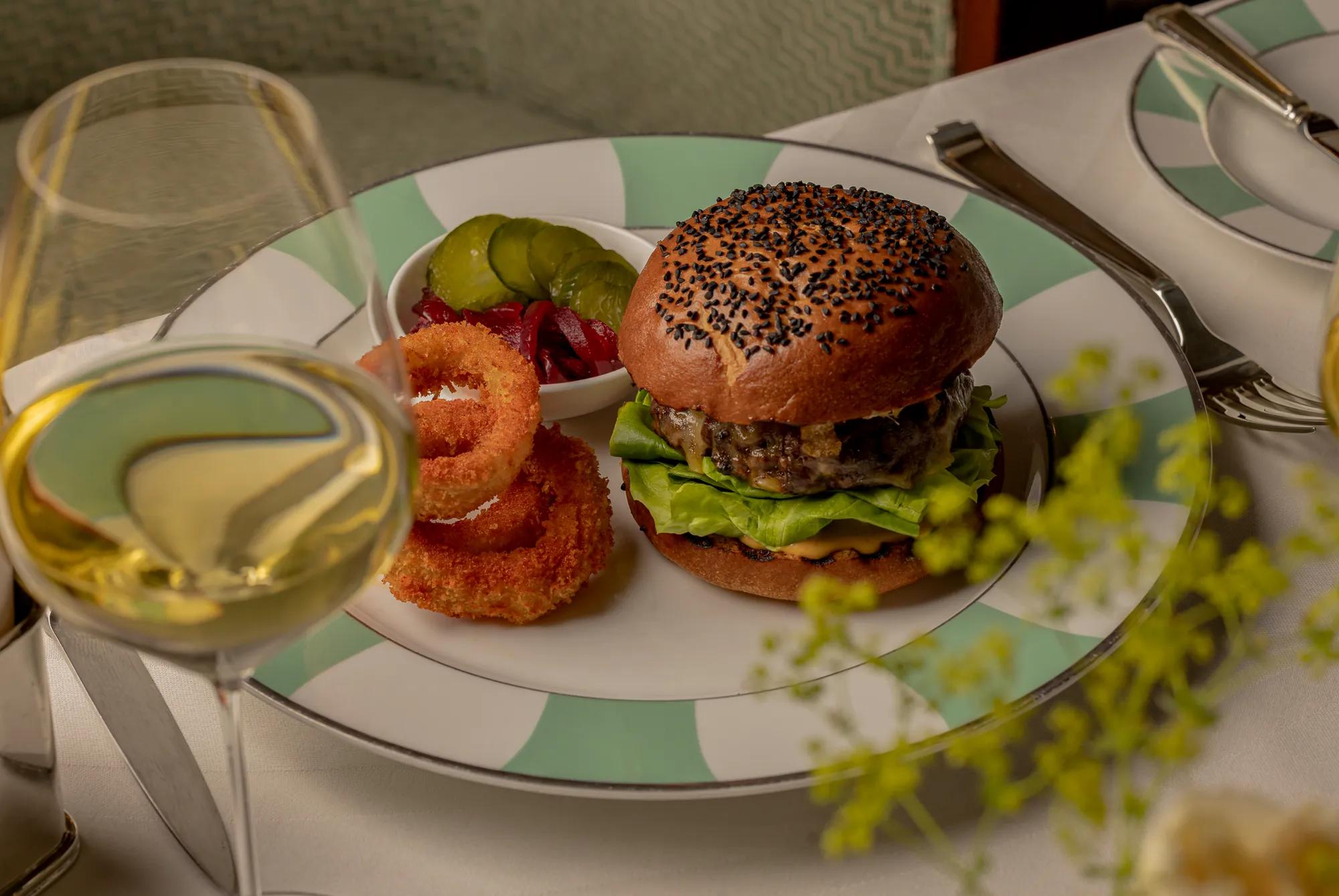 Gourmet burger served on a sesame-topped bun with lettuce and sauce, plated alongside crispy onion rings, pickles, and red onions. Presented on green-and-white patterned china with a glass of white wine on a neatly set table.