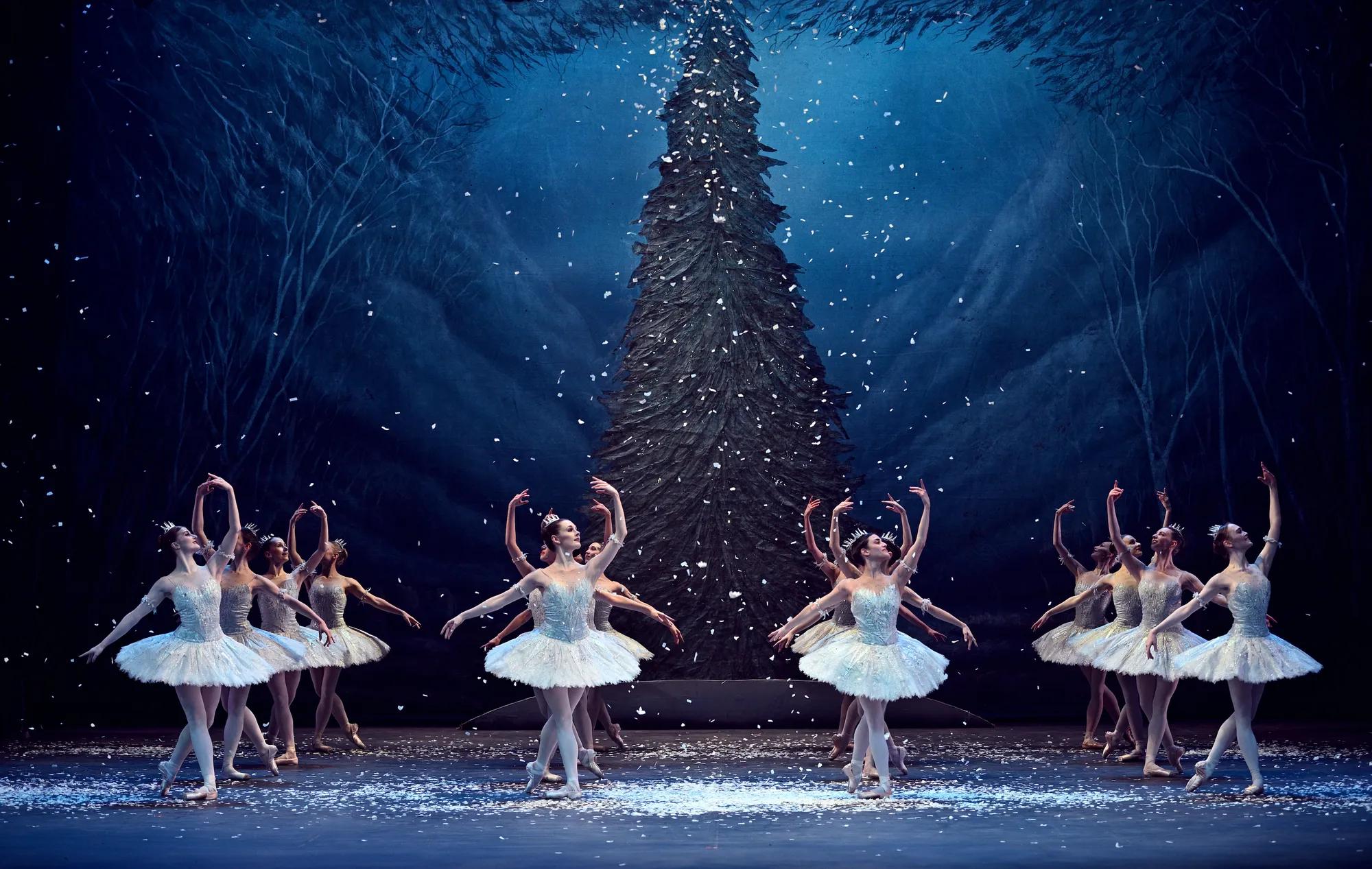 Ballet dancers in white tutus performing amid falling snow on a winter forest stage.