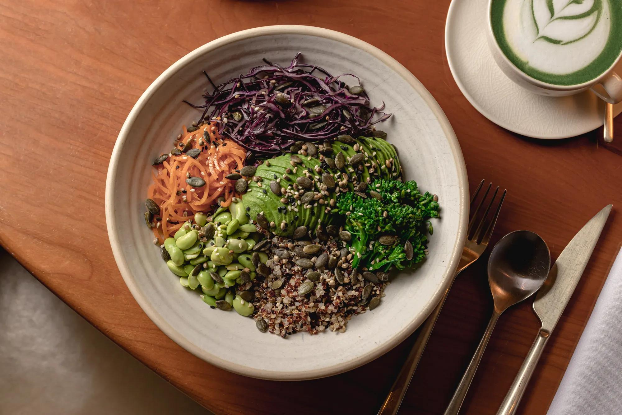 A healthy, vibrant bowl filled with shredded carrots, purple cabbage, avocado slices, edamame, broccoli, quinoa, and pumpkin seeds, arranged in sections on a beige plate. A fork and spoon are placed next to the plate on a wooden table.