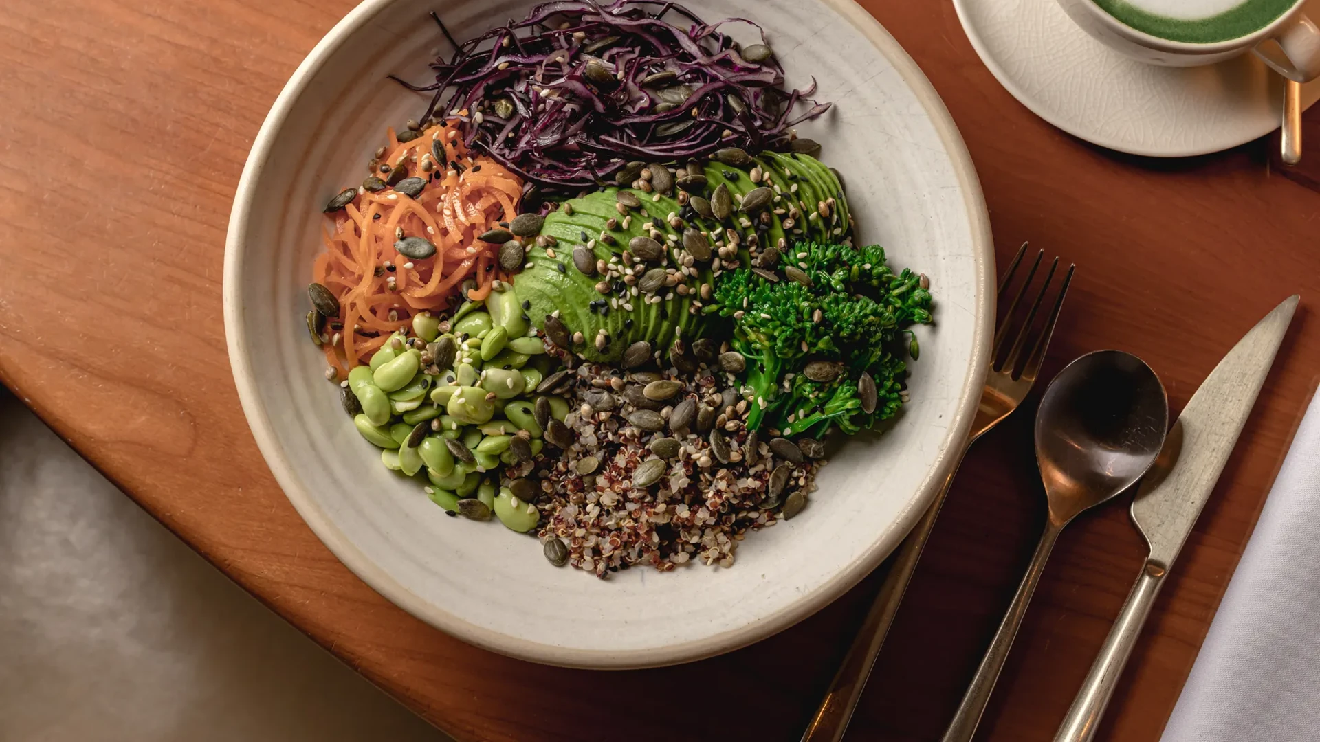 A healthy, vibrant bowl filled with shredded carrots, purple cabbage, avocado slices, edamame, broccoli, quinoa, and pumpkin seeds, arranged in sections on a beige plate. A fork and spoon are placed next to the plate on a wooden table.