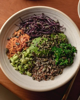 A healthy, vibrant bowl filled with shredded carrots, purple cabbage, avocado slices, edamame, broccoli, quinoa, and pumpkin seeds, arranged in sections on a beige plate. A fork and spoon are placed next to the plate on a wooden table.