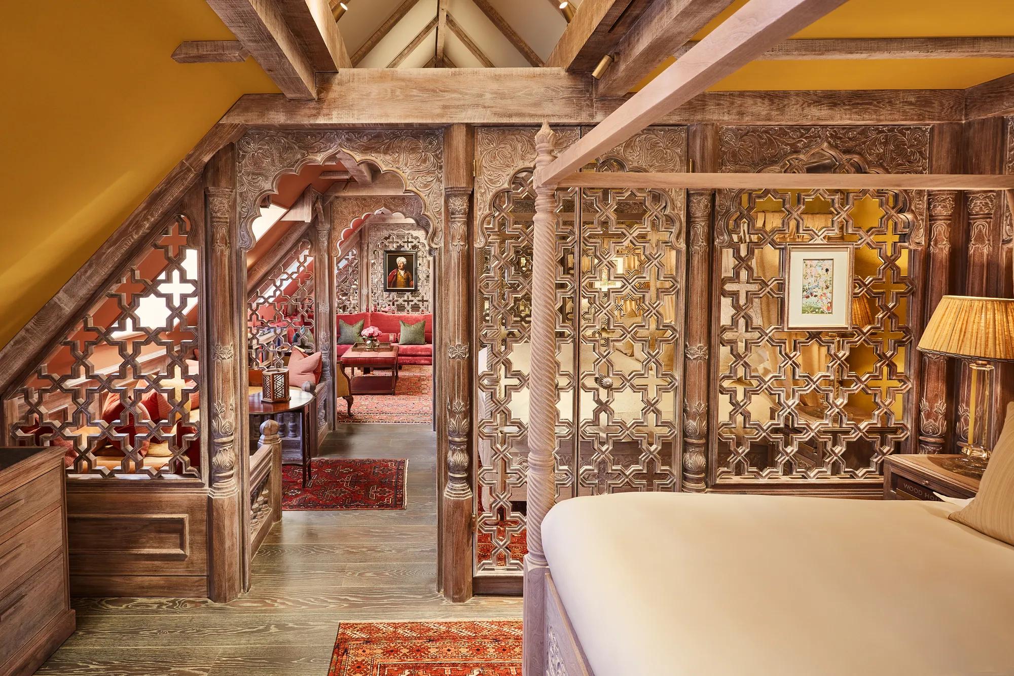 Ornate wood-carved lounge with red seating, patterned rug, small table, and leaded window under sloped ceiling.