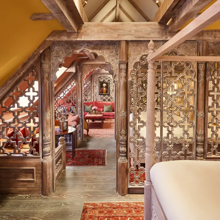Ornate wood-carved lounge with red seating, patterned rug, small table, and leaded window under sloped ceiling.