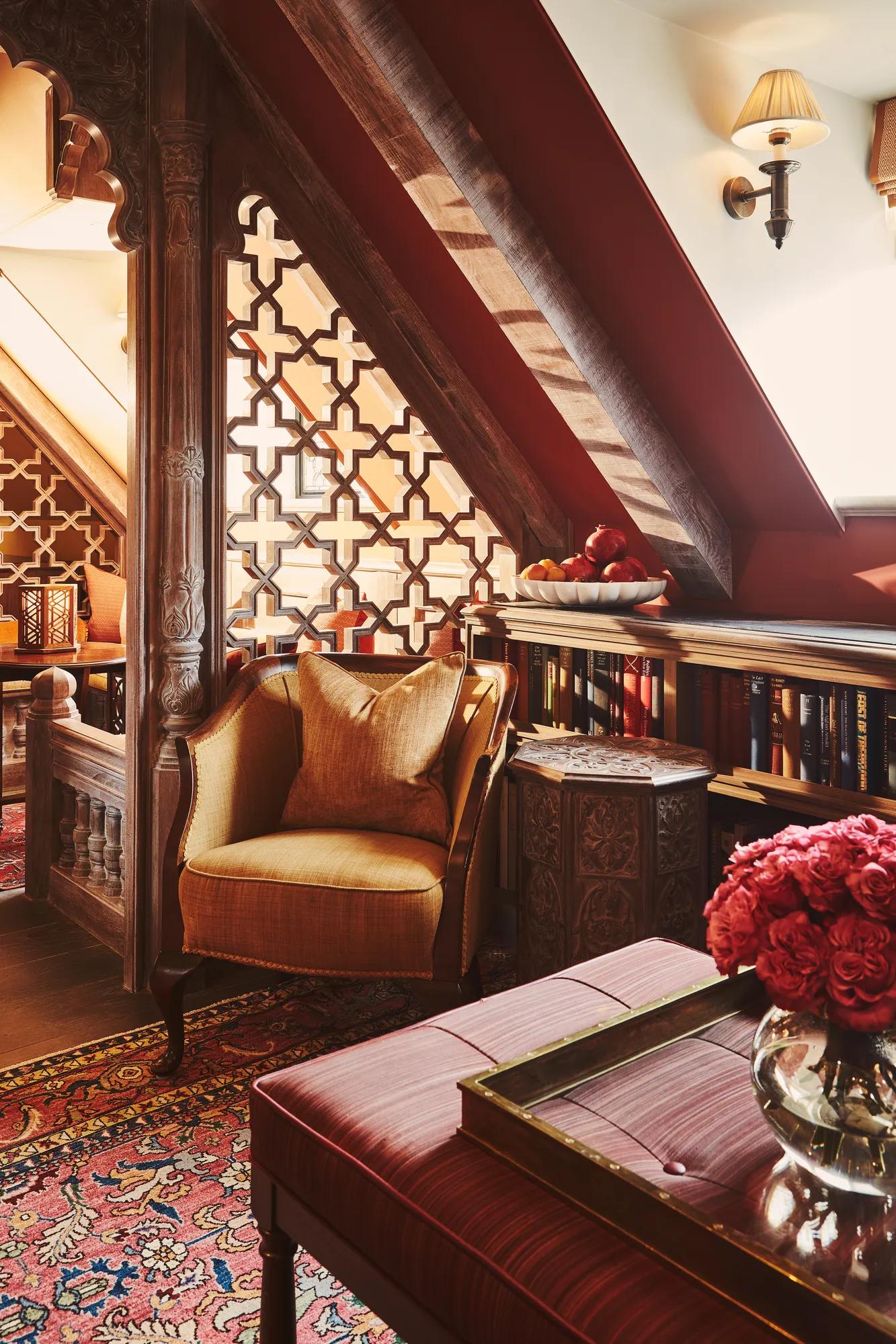 Ornate lounge with carved wood screens, patterned rug, upholstered chair, bookshelves, and flowers beneath sloped ceiling and wall sconce.