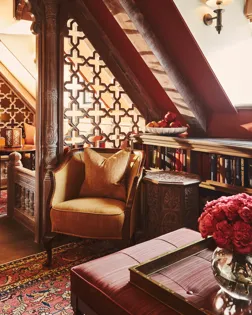 Ornate lounge with carved wood screens, patterned rug, upholstered chair, bookshelves, and flowers beneath sloped ceiling and wall sconce.