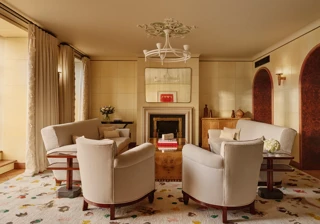 Warm, elegant living room with cream armchairs and sofas arranged around a wooden coffee table and fireplace, featuring arched doorways, patterned rug, and soft natural light through tall curtains.