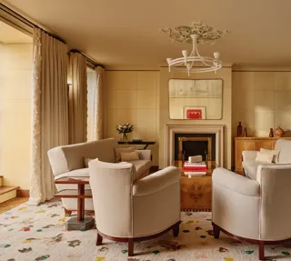 Warm, elegant living room with cream armchairs and sofas arranged around a wooden coffee table and fireplace, featuring arched doorways, patterned rug, and soft natural light through tall curtains.