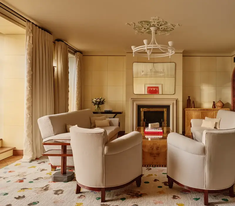 Warm, elegant living room with cream armchairs and sofas arranged around a wooden coffee table and fireplace, featuring arched doorways, patterned rug, and soft natural light through tall curtains.