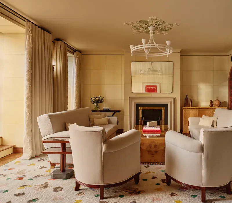Warm, elegant living room with cream armchairs and sofas arranged around a wooden coffee table and fireplace, featuring arched doorways, patterned rug, and soft natural light through tall curtains.