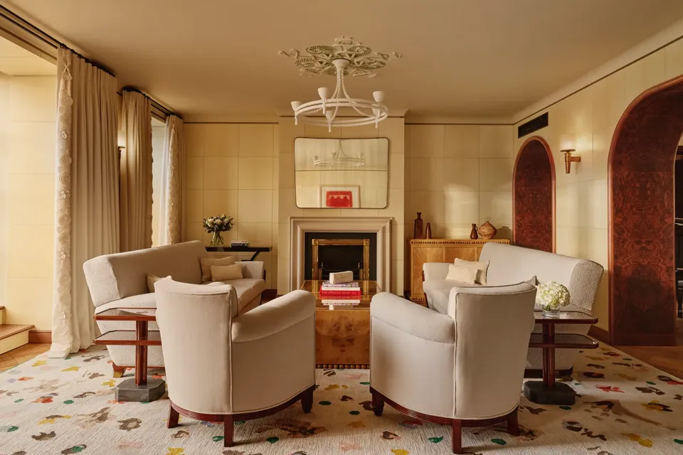 Warm, elegant living room with cream armchairs and sofas arranged around a wooden coffee table and fireplace, featuring arched doorways, patterned rug, and soft natural light through tall curtains.