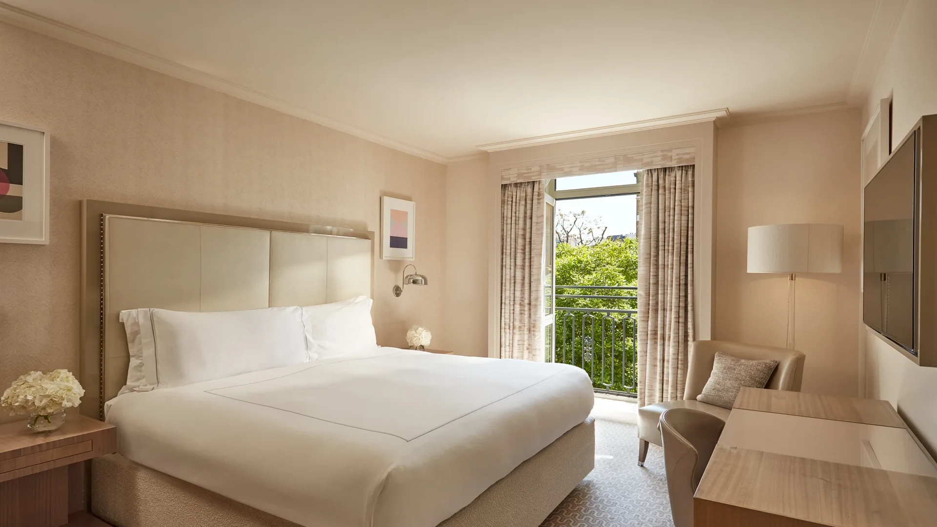 Neutral-toned Belgravia Room with king bed, desk, armchair, and French doors opening to a balcony with greenery.