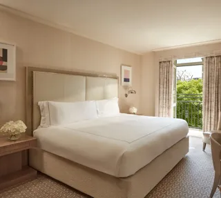 Neutral-toned Belgravia Room with king bed, desk, armchair, and French doors opening to a balcony with greenery.