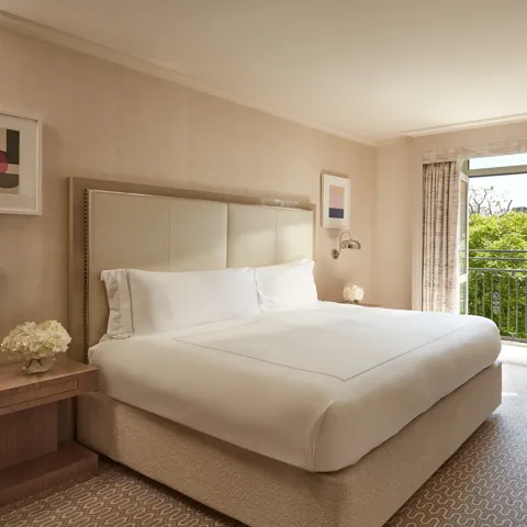 Neutral-toned Belgravia Room with king bed, desk, armchair, and French doors opening to a balcony with greenery.