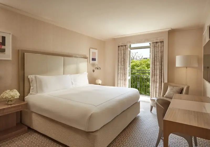 Neutral-toned Belgravia Room with king bed, desk, armchair, and French doors opening to a balcony with greenery.
