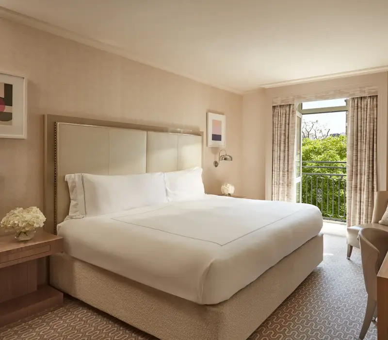 Neutral-toned Belgravia Room with king bed, desk, armchair, and French doors opening to a balcony with greenery.