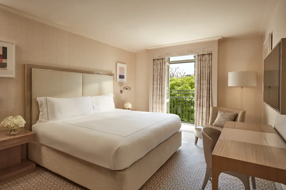 Neutral-toned Belgravia Room with king bed, desk, armchair, and French doors opening to a balcony with greenery.