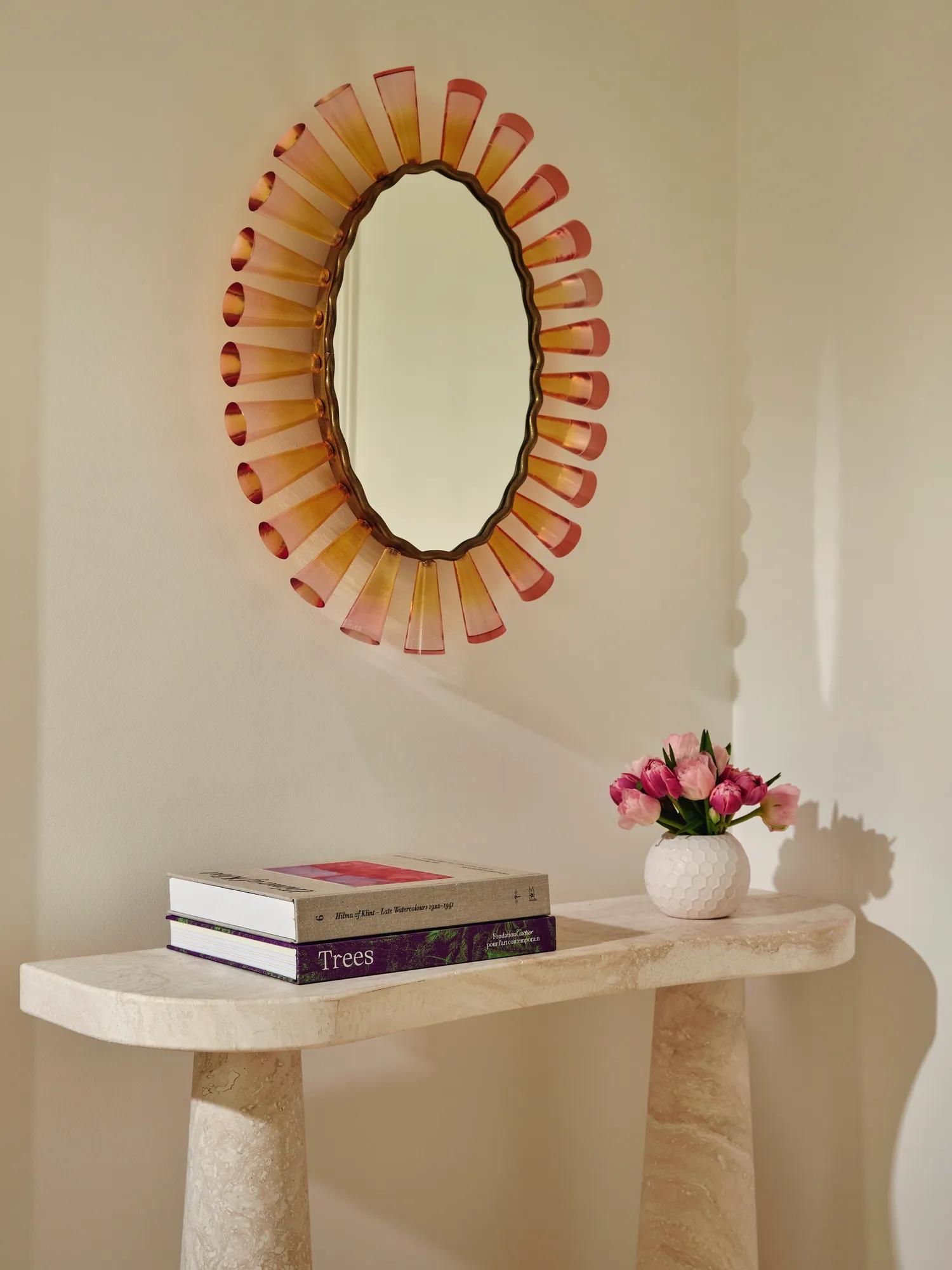 Stone console with art books and pink flowers beneath an oval mirror framed in pink glass.