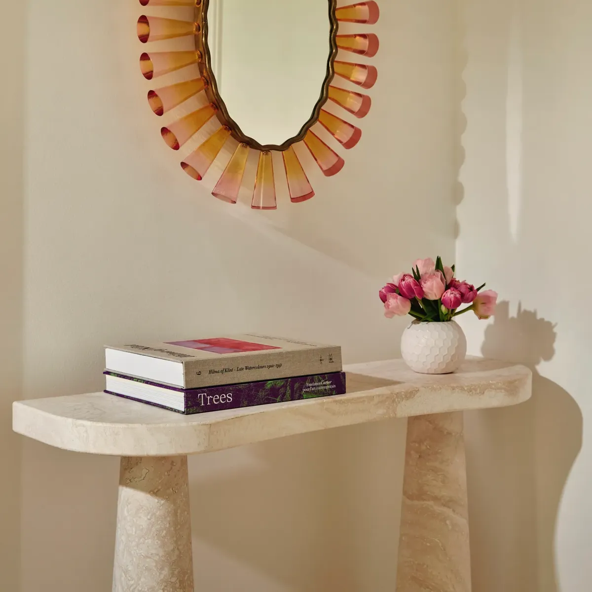 Stone console with art books and pink flowers beneath an oval mirror framed in pink glass.