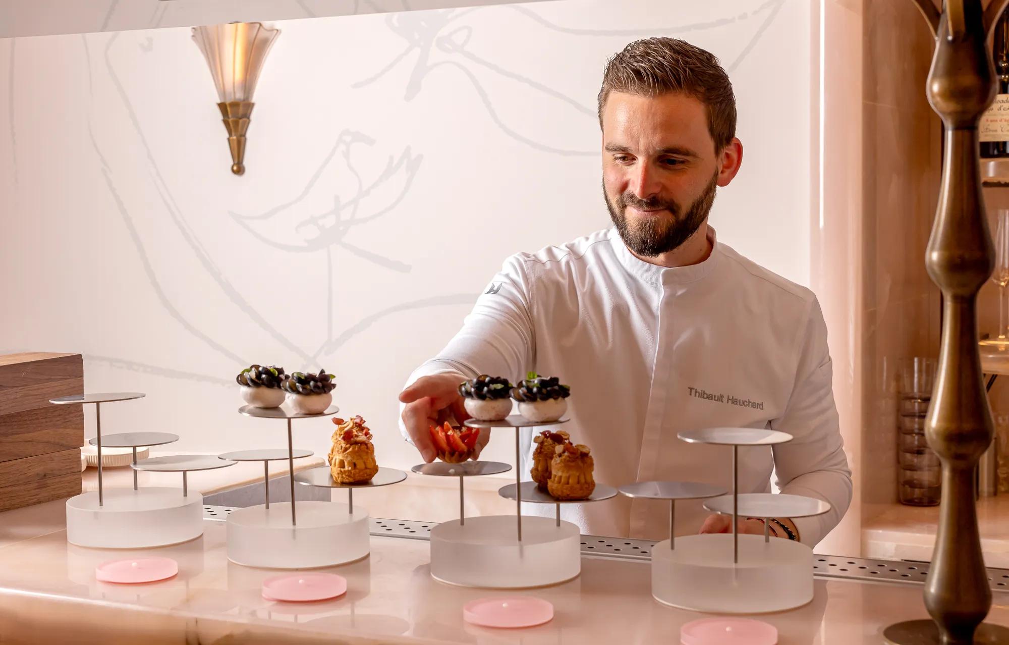 A pastry chef wearing a white chef’s jacket carefully arranges elegant desserts topped with berries and cream on tiered stands at a marble counter in a softly lit patisserie setting.