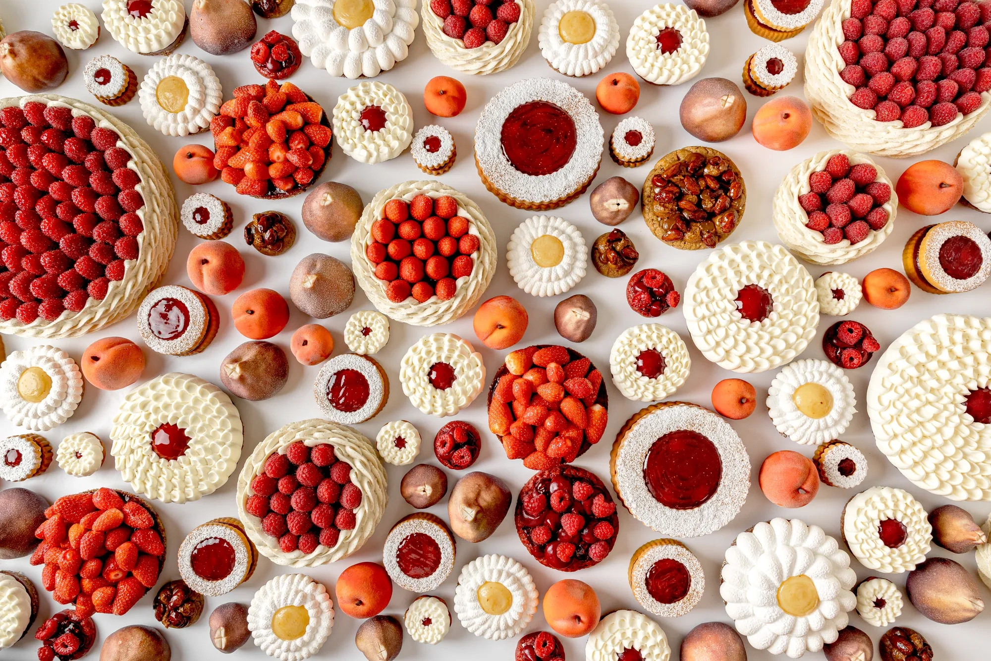 An overhead view of an artistic arrangement of fruit tarts and cakes topped with raspberries, strawberries, cream, and fruit glazes, displayed alongside whole peaches and figs on a white background.