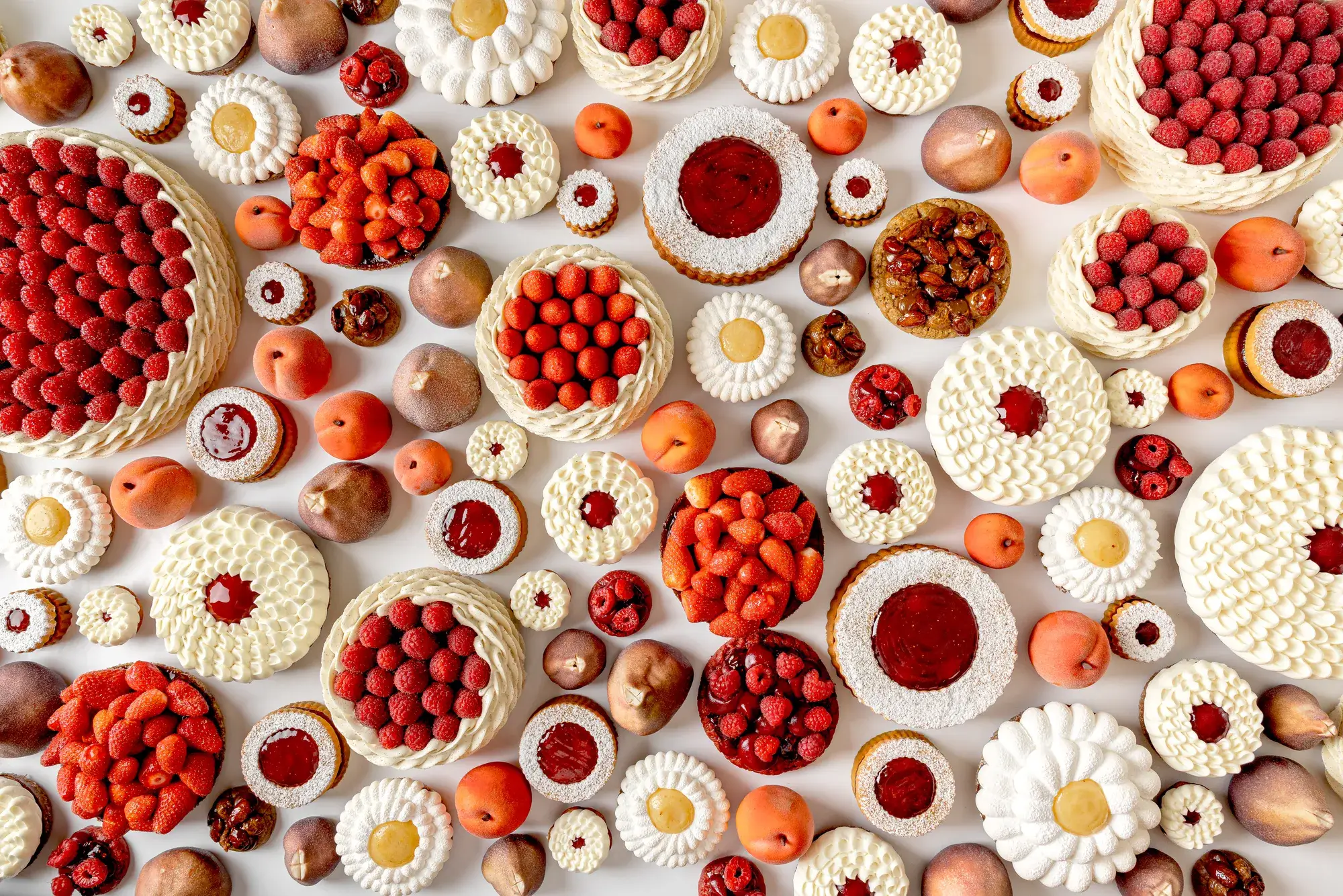 An overhead view of an artistic arrangement of fruit tarts and cakes topped with raspberries, strawberries, cream, and fruit glazes, displayed alongside whole peaches and figs on a white background.