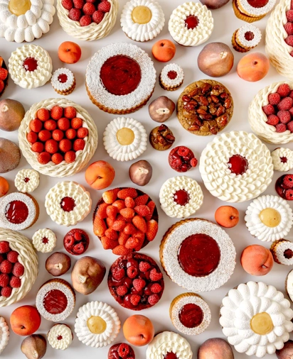 An overhead view of an artistic arrangement of fruit tarts and cakes topped with raspberries, strawberries, cream, and fruit glazes, displayed alongside whole peaches and figs on a white background.