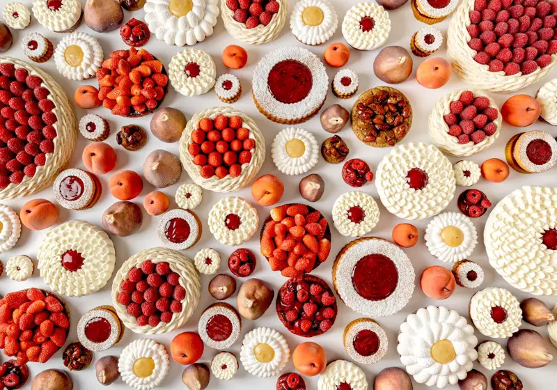 An overhead view of an artistic arrangement of fruit tarts and cakes topped with raspberries, strawberries, cream, and fruit glazes, displayed alongside whole peaches and figs on a white background.