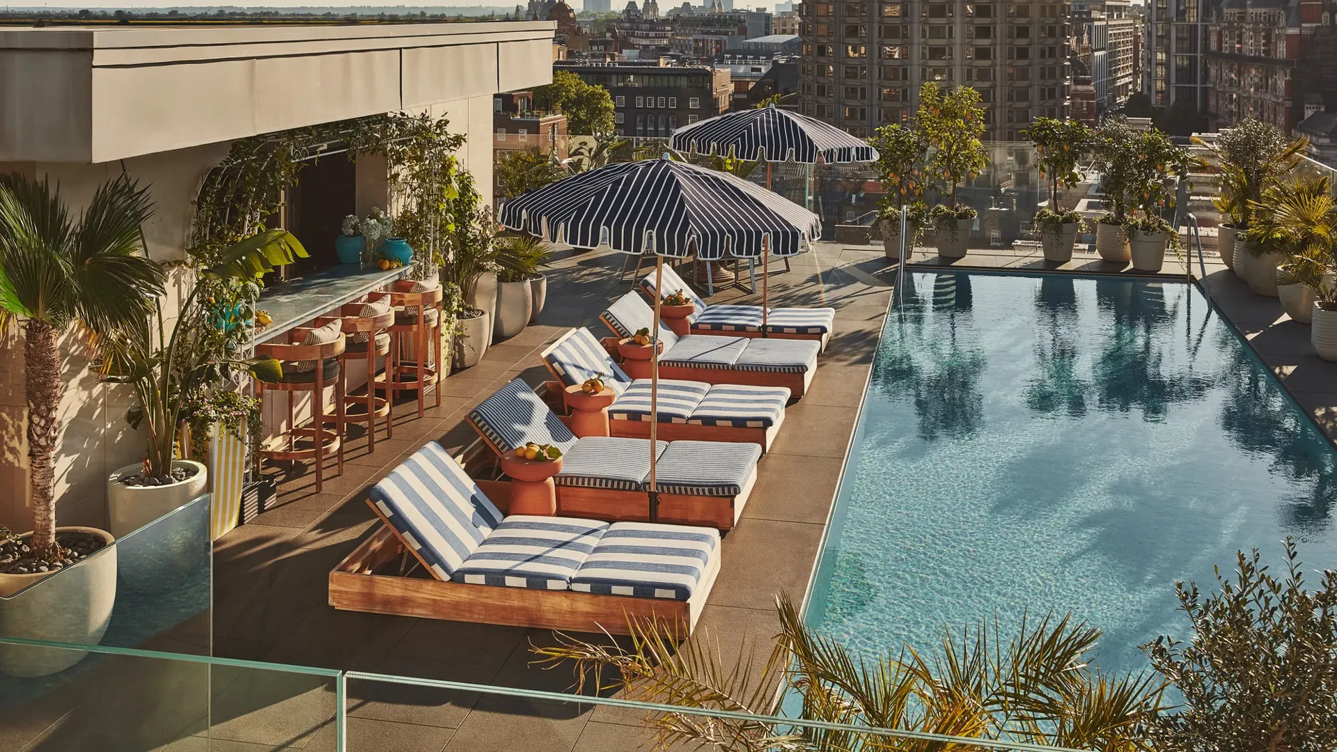 Rooftop pool with striped loungers, bar seating, greenery, and city skyline views.