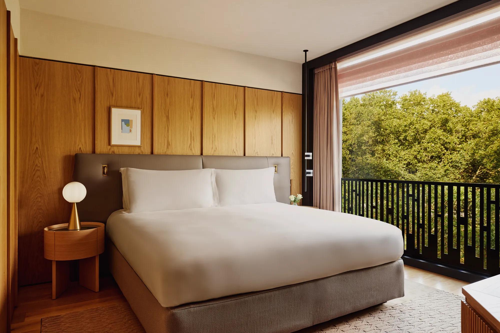 Hotel bedroom with king bed, bedside lamps, and city view through large window.