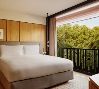 Hotel bedroom with king bed, bedside lamps, and city view through large window.