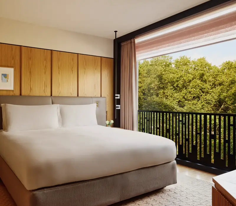 Hotel bedroom with king bed, bedside lamps, and city view through large window.