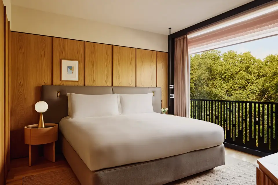 Hotel bedroom with king bed, bedside lamps, and city view through large window.