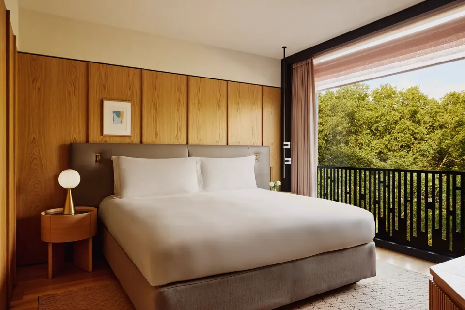 Hotel bedroom with king bed, bedside lamps, and city view through large window.