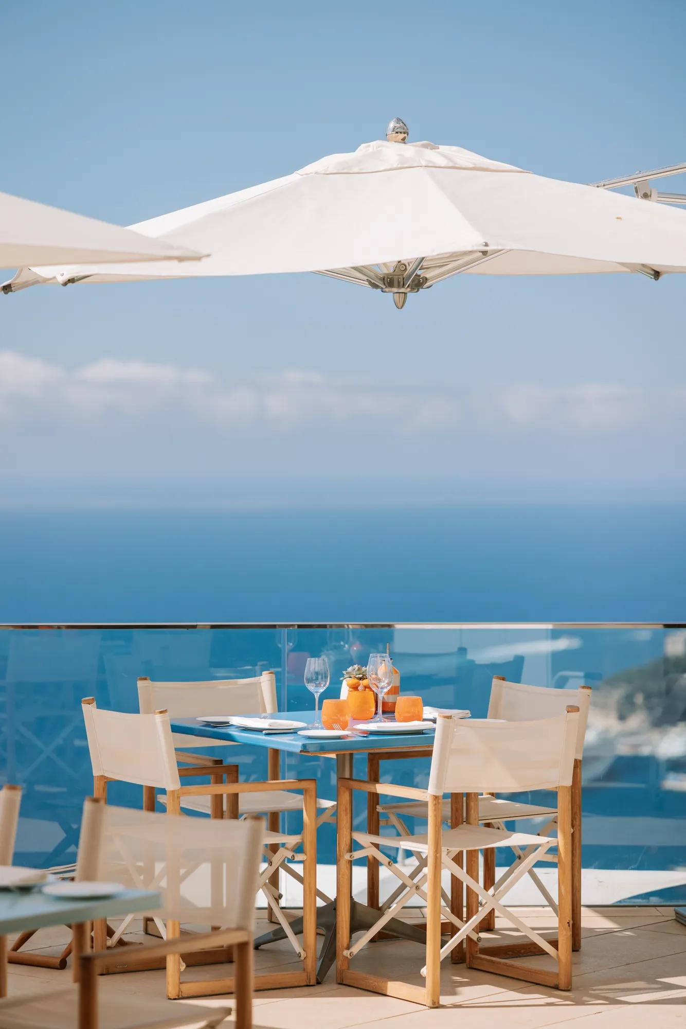 Table de repas en extérieur dressée avec des boissons sous un parasol blanc, offrant une vue panoramique sur la mer depuis une terrasse vitrée.