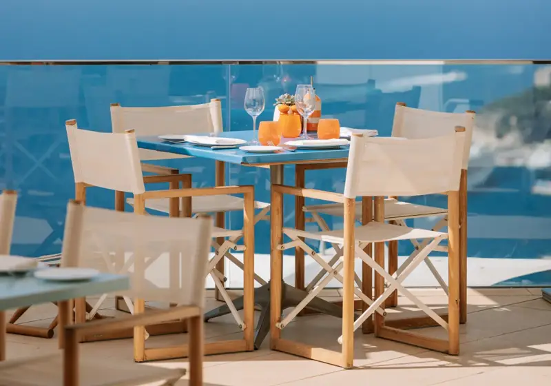 Table de repas en extérieur dressée avec des boissons sous un parasol blanc, offrant une vue panoramique sur la mer depuis une terrasse vitrée.