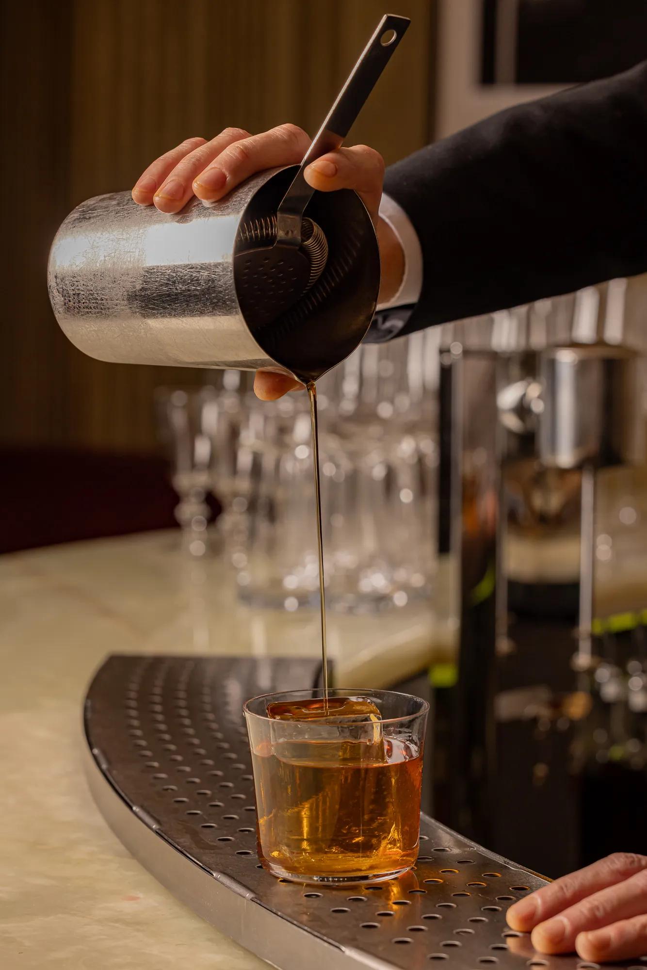Bartender pouring an amber cocktail from a metal shaker through a strainer into a glass with a large ice cube on a marble bar counter.