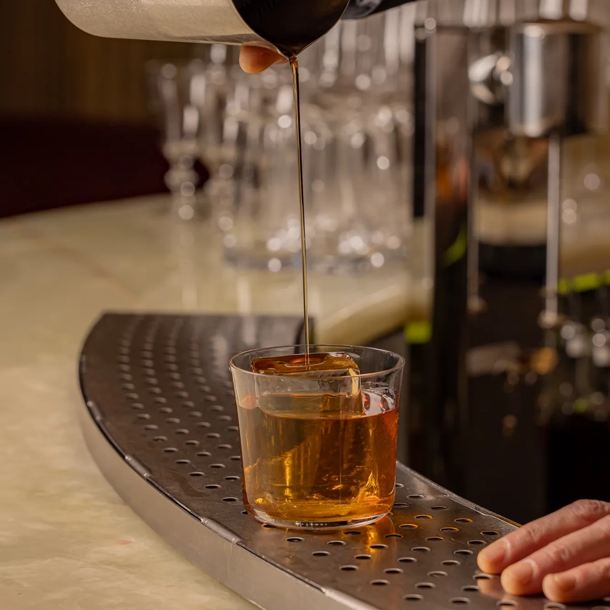 Bartender pouring an amber cocktail from a metal shaker through a strainer into a glass with a large ice cube on a marble bar counter.