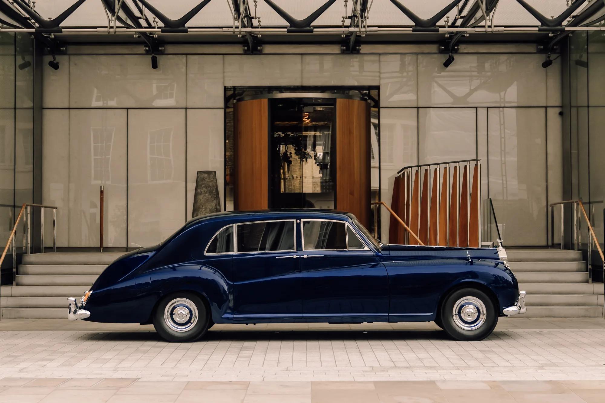 A classic dark blue luxury car is parked in front of a grand hotel entrance with a revolving wooden door and wide stone steps on either side.
