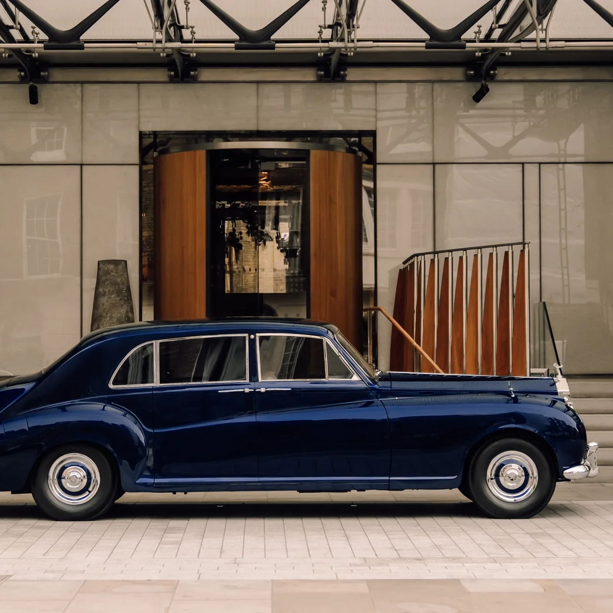 A classic dark blue luxury car is parked in front of a grand hotel entrance with a revolving wooden door and wide stone steps on either side.